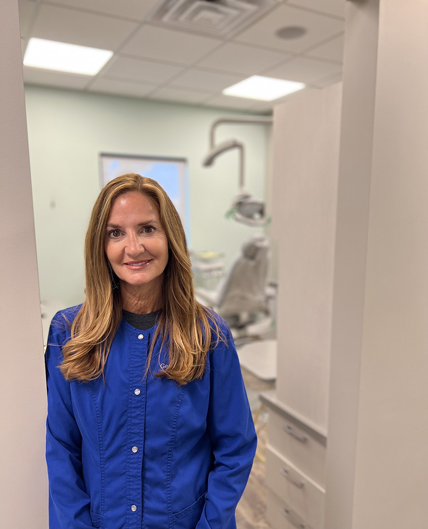 The image shows a woman standing in an indoor setting, likely a dental or medical office, wearing a blue shirt and jacket, with a smile on her face, posing for the camera.