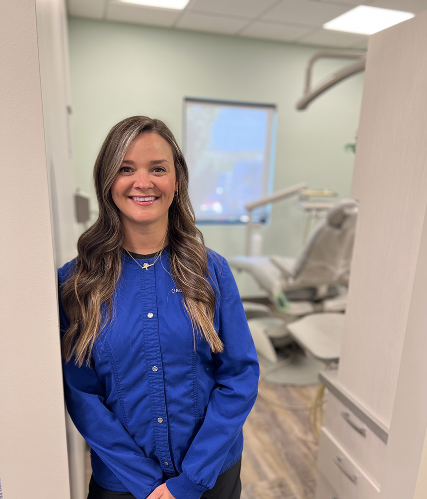 Woman standing in front of dental office equipment with a blue shirt and long hair.