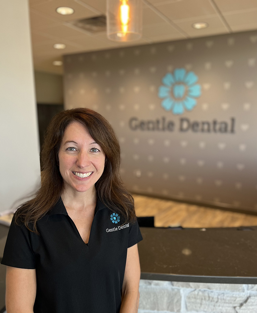 The image shows a woman standing in front of a sign with the text  Gentle Dental  on it, posing for a photograph.