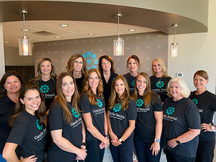 The image shows a group of individuals wearing black t-shirts with white text and logos, posing together for a photograph in an indoor setting with a modern decor.