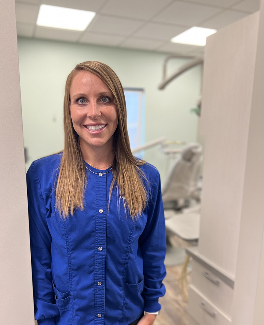 A woman wearing a blue shirt stands in front of a dental office chair with a smiling expression, positioned between two walls with visible tile patterns.