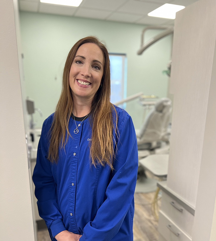 Woman standing in dental office with smiling expression, wearing blue jacket and name tag.