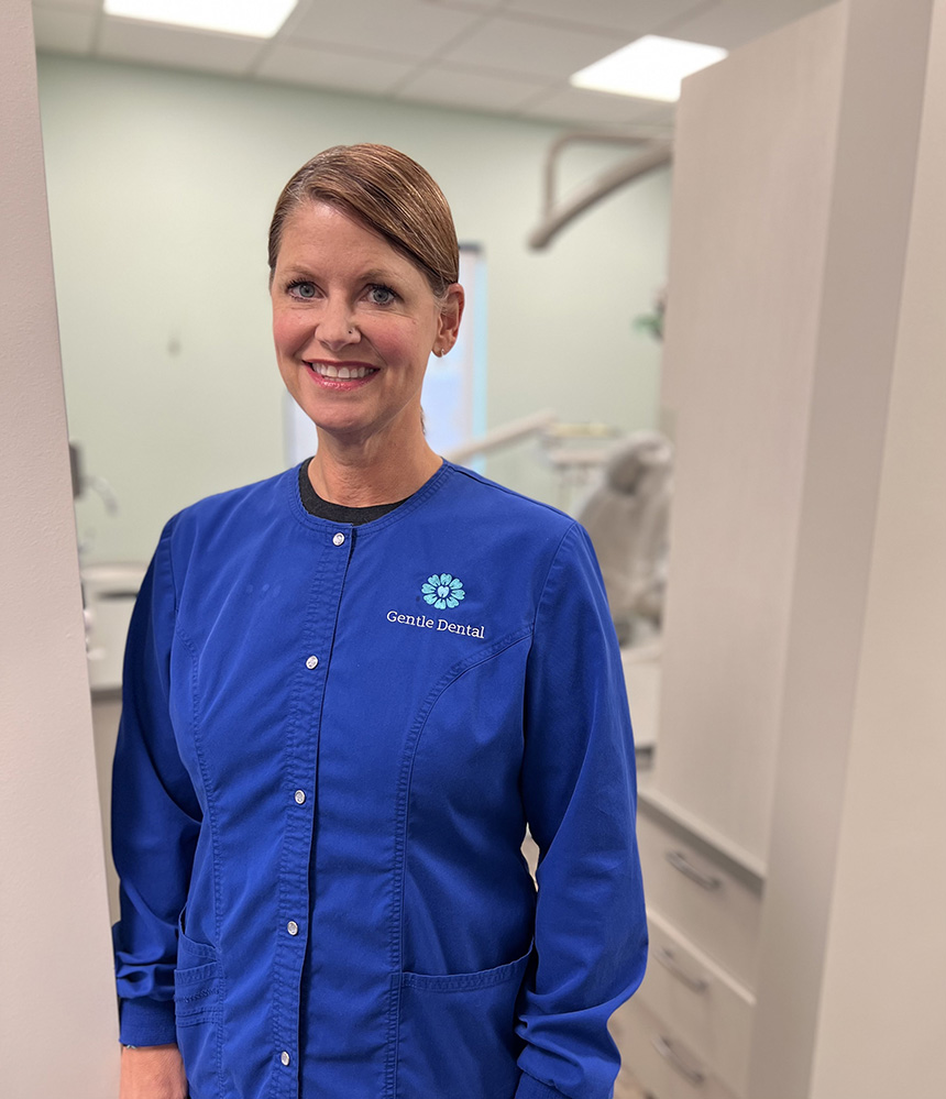 A woman wearing a blue coat stands in front of a dental office chair.
