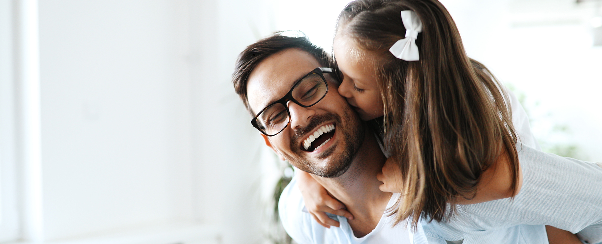 The image shows a family of three, including an adult couple and a child, with the adults smiling and embracing each other while looking at the camera, set against a blurred background that suggests an indoor setting.