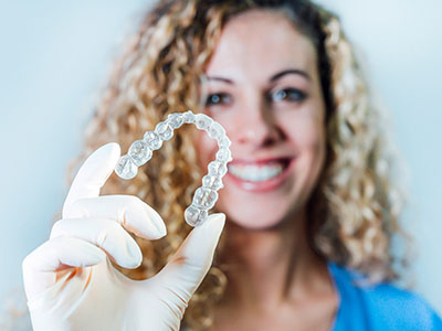 The image shows a woman holding up a clear dental retainer with her hand.