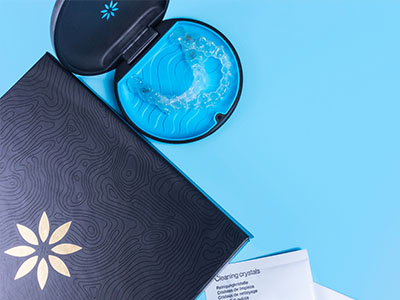 The image shows a tabletop with an open box containing a blue object resembling a small, round, decorative item, possibly a paperweight, next to a cup of water with a floating flower, and a closed gift box with a patterned design and a logo on the lid.