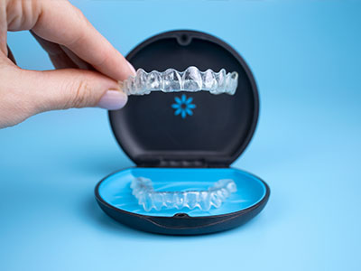 The image shows a person s hand holding a clear plastic tray containing a set of transparent aligners, with one aligner placed inside a black case on a blue surface.