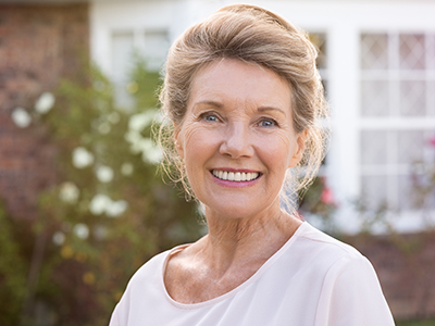 The image shows an older woman with short hair wearing a light-colored top, posing outdoors with a smile against a backdrop of a house and garden.