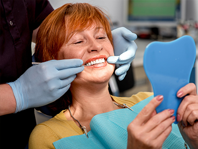 A woman with red hair sitting in a dental chair, smiling at the camera while holding up a blue toothbrush, with dental professionals attending to her.