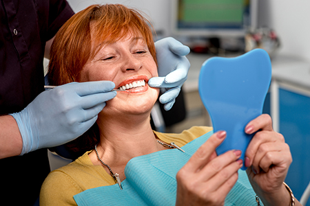 A woman sitting in a dental chair with a blue smiley face mask on her teeth, holding a mirror while a dental professional works on her teeth.