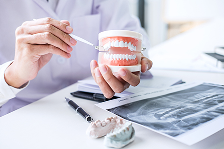 A dentist holding a dental model with a hand tool, examining it closely while seated at a workstation with various dental tools and models around.