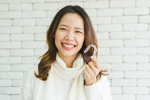 A woman with a smile, holding a small object close to her face, poses for a portrait against a brick wall background.