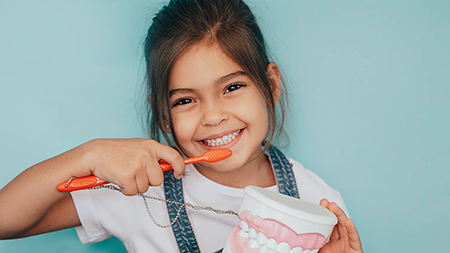 The image features a young girl brushing her teeth with an oversized toothbrush, holding a toothpaste tube, while standing against a blue background.