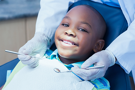 A young boy with a big smile sitting in a dental chair, being attended to by a dentist wearing gloves and a mask.