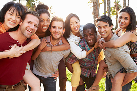 A group of young adults posing together outdoors with smiles on their faces, embracing each other for a photo, under a bright sky.