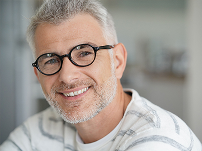 The image shows a man with glasses smiling at the camera, appearing to be middle-aged, with grey hair and a beard, wearing a striped shirt. He has a friendly expression and appears to be sitting indoors.