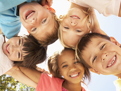 The image shows a group of six children posing together with smiles on their faces, looking directly at the camera, with a clear blue sky in the background.