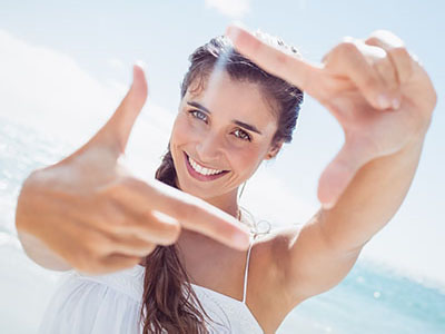 The image shows a young woman taking a selfie with her hand held out towards the camera, smiling at the camera, with a bright, sunny background suggesting a beach setting.
